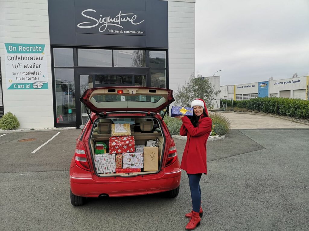 Véronika avec un cadeau dans les mains devant SignaturePub avec les cadeaux de Noël pour les Boîtes Solidaires